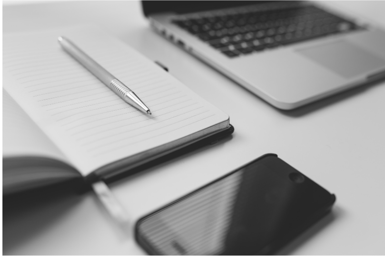 black and white office materials. Laptop, cellphone, pen, a notebook and a mouse.